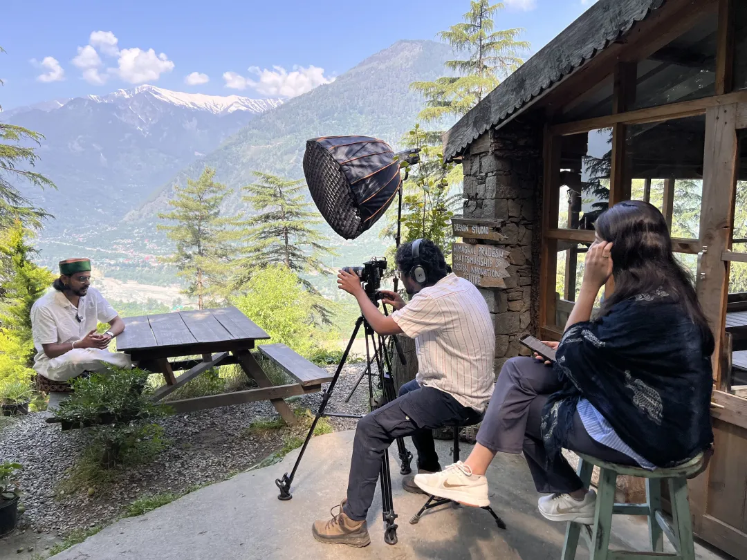 A man sitting at a park bench in front of a camper for an interview; mountains form a majestic background.